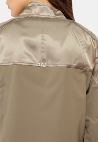 Giacca kaki con una spallina in satin beige lucido. Caratterizzata da un distintivo gancio in metallo e una texture liscia.