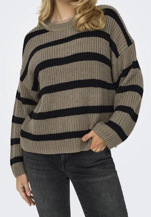 Personne portant un pull en maille taupe avec des rayures horizontales noires et un jean gris foncé, une main dans la poche, sur un fond clair uni.