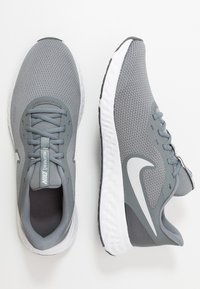 Šedé atletické boty vyrobené z prodyšného materiálu, s polstrovaným jazykem, šedými tkaničkami a bílou gumovou podrážkou s texturovaným vzorem.