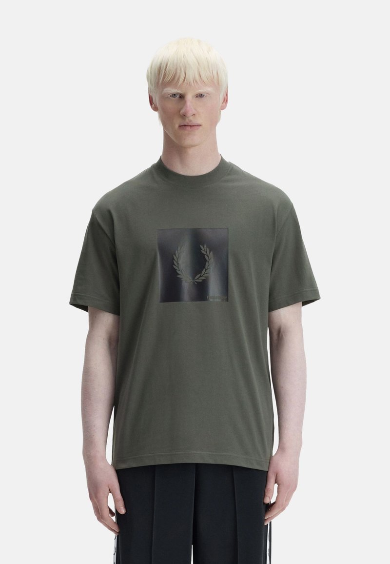 Jeune personne aux cheveux pâles porte un T-shirt vert olive avec un carré noir brillant et un logo de couronne de laurier sur la poitrine.