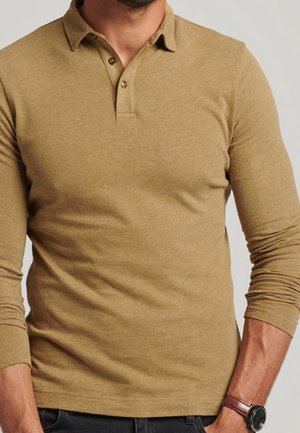 Mężczyzna w dopasowanej, beżowej koszulce polo z długim rękawem, z dwoma guzikami i zegarkiem na lewym nadgarstku.