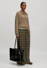 Bruine gebreide polo trui met drie knopen, gecombineerd met groene geruite wijde broek. Zwarte glanzende schoudertas in de hand, beige schoenen.