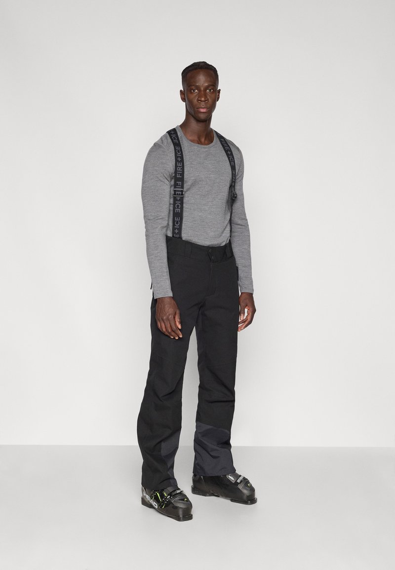 Μαύροι σκι παντελόνια με ρυθμιζόμενους μαύρους ιμάντες, γκρι μακρυμάνικο πουκάμισο και μαύρα παπούτσια του σκι με πράσινες λεπτομέρειες, σε λευκό φόντο.