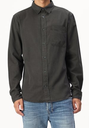 Homme portant une chemise à manches longues boutonnée gris foncé avec poche poitrine et un jean bleu délavé, debout devant un fond blanc.