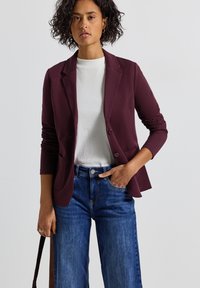 Blazer bordeaux con rever e due bottoni, indossato su un maglione bianco a coste. Abbinato a jeans a gamba larga blu. Texture liscia.