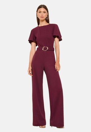 Femme portant une combinaison bordeaux avec des manches papillon et une ceinture à la taille ornée d'une boucle géométrique dorée, debout devant un fond uni.