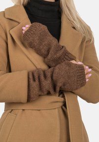 Guantes de punto marrones sin dedos, usados sobre un cuello de tortuga negro, combinados con un abrigo color camel con solapas anchas y una cintura ceñida con cinturón.