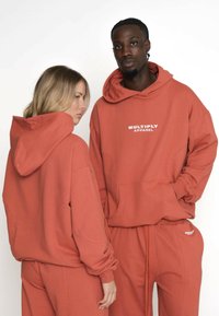 Două modele purtând hanorace și pantaloni de trening de culoare ruginie, unul întors cu fața spre cameră și celălalt ușor întors.
