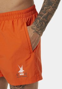 Pantaloni da bagno arancioni con elastico in vita, tasca laterale e logo Helly Hansen bianco sull'orlo. Tessuto liscio e leggero.
