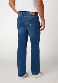 Mėlyni džinsai su tiesiu kirpimu, turintys odinį logotipo ženkliuką ant juosmens ir dviems užpakalinėms kišenėms su trikampiais akcentais.