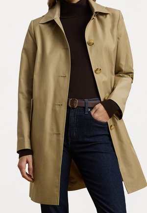 Personne portant un trench-coat beige sur un pull à col roulé noir, un jean bleu foncé et une ceinture marron, une main dans la poche.
