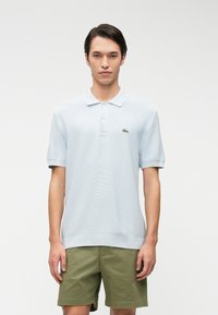 Polo shirt bleu clair à manches courtes avec un design texturé, muni d'un plastron à deux boutons et d'un logo brodé vert sur la poitrine.