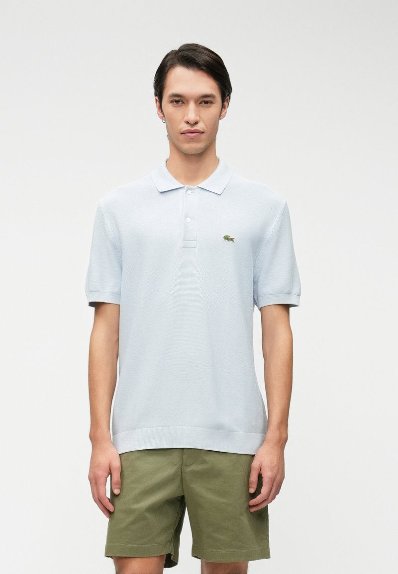 Polo shirt bleu clair à manches courtes avec un design texturé, muni d'un plastron à deux boutons et d'un logo brodé vert sur la poitrine.