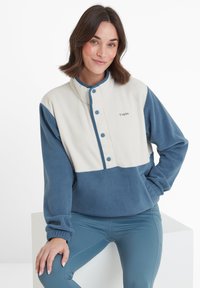 Woman with shoulder-length brown hair wearing a blue and white fleece pullover and blue leggings, seated with a relaxed pose and slight smile.