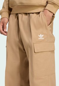 Pantalones cargo beige con una tela texturizada, que presentan bolsillos laterales, una cinturilla elástica y un logo blanco de Adidas en la parte frontal.