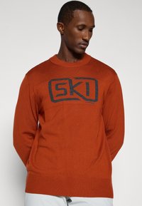 Oranžna pletena jopica z okroglim ovratnikom in rebrastimi manšeti. Ponuja izrazit črn logo "SKI" spredaj, gladka tekstura.