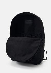 Sac à dos noir avec compartiment principal ouvert, poche avant zippée et étiquette intérieure visible sur fond clair uni.