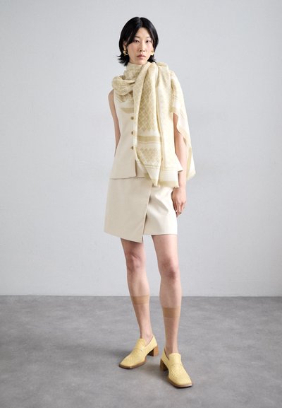 Haut sans manches beige avec des boutons, jupe assortie à ourlet asymétrique et talons jaunes texturés. Une écharpe jaune à motifs drape les épaules.