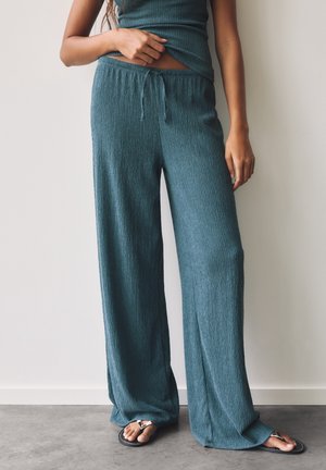 Femme portant un pantalon large bleu sarcelle et un haut assorti, debout pieds nus sur un sol en béton contre un mur blanc.