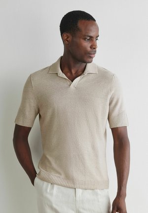 Młody mężczyzna z krótkimi włosami, ubrany w beżowy polo z krótkim rękawem i jasne spodnie, patrzący w prawo na tle jednolitej ściany.