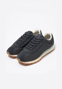 Zapatillas deportivas gris oscuro que presentan una mezcla de materiales lisos y texturizados, un elemento de diseño dentado y una suela beige con un patrón de olas.