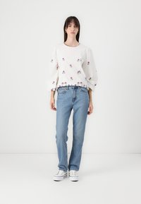 Blusa bianca con ricami floreali viola, maniche a sbuffo e scollo a scallop, abbinata a jeans blu chiaro a gamba dritta e sneakers bianche.