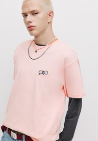 Tricou roz deschis cu guler rotund, mâneci scurte și un design brodat cu ochelari triști pe piept. Suprapus cu o cămașă lungă de culoare închisă dedesubt.