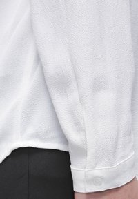 Vit texturerad blus med knappärmar, som har långa ärmar och en rundad nederkant, visad mot en svart bakgrund.