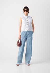 Colete acolchoado branco com botões dourados, jeans de perna larga azul-claro, óculos de sol castanhos e uma pequena mala bordô. Textura e ajuste destacados.