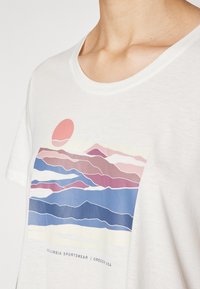 Bijela majica kratkih rukava s grafikom šarenog planinskog krajolika u ružičastoj, plavoj i krem boji, s logotipom Columbia Sportswear ispod.