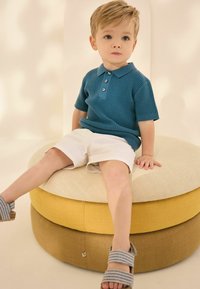 Tyrkysový žebrovaný polo trikoty s třemi knoflíky, nošený s bílými šortkami, sedící na kulatém, vrstveném, vícebarevném potahu.