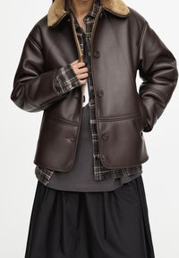 Veste en cuir marron avec un col en fourrure beige, dotée d'une fermeture à boutons et de deux poches latérales. Portée par-dessus une chemise à carreaux et une jupe noire.