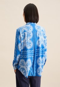 Blusa di seta blu con motivi paisley bianchi, maniche lunghe e design con piega sul retro. Tessuto morbido e vestibilità comoda.