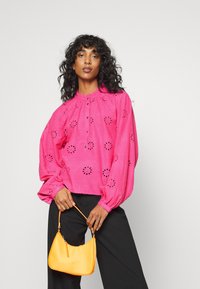 Blusa rosa de mangas longas com recortes florais, decote franzido e fecho de botões, combinada com uma pequena bolsa amarela. Textura lisa.