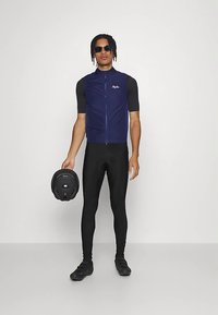 Vyras, vilkintis juodas dviratininko kelnes, juodą trumpą rankovę marškinėlį, tamsiai mėlyną pasišalintą liemenę su užtrauktuku, saulės akinius ir laikančius juodą dviratininko šalmą.