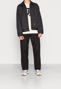 Person klädd i svart jacka, beige grafisk t-shirt, svarta byxor och teal-vita sneakers stående på en enkel ljusgrå golv mot vit bakgrund.