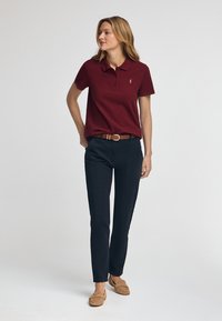 Bordeaux polo shirt met korte mouwen, kraag en klein logo; gecombineerd met strakke marineblauwe broek en tan moccasin-stijl schoenen.