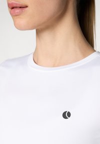 Chemise blanche ajustée avec une texture lisse, ornée d'un petit logo noir sur la poitrine. Col rond et design minimaliste.