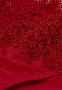 Rotes Kleidungsstück mit einem Spitzenüberzug, der komplizierte Blumenmuster im oberen Bereich aufweist, ergänzt durch eine glatte, einfarbige rote Basis.