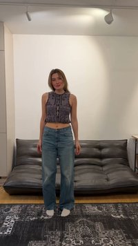 Junge Frau trägt ein gemustertes ärmelloses Oberteil und blaue Jeans, steht barfuß auf einem gemusterten Teppich in einem modernen Wohnzimmer.