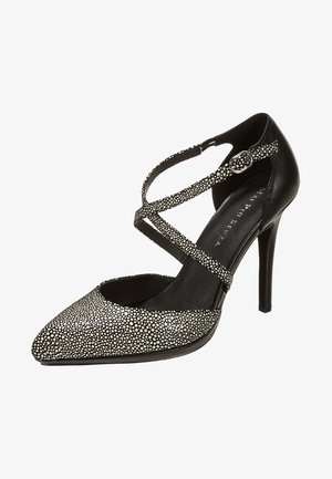 Zapato de tacón alto negro con punta afilada salpicada de blanco y correas cruzadas en el tobillo aseguradas por una pequeña hebilla de plata.