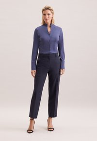 Satijnen blauwe blouse met een hoge kraag en knoopsluiting, gecombineerd met op maat gemaakte marineblauwe broek en zwarte sandalen met bandjes. Over het geheel een gladde textuur.