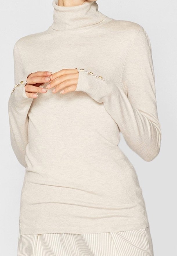 Pull beige à col roulé avec détails de boutons sur les poignets, porté avec un pantalon rayé de couleur claire, vu du cou jusqu'aux hanches.