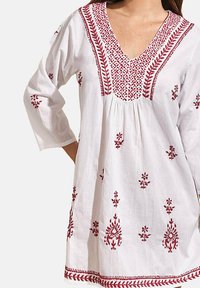Tunique blanche ornée de broderies florales rouges, avec un décolleté en V profond et des manches trois-quarts. Le tissu est léger et légèrement texturé.