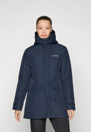 Vrouw in een marineblauwe waterdichte Didriksonsjas met hoge kraag en capuchon, staand tegen een effen lichte achtergrond.