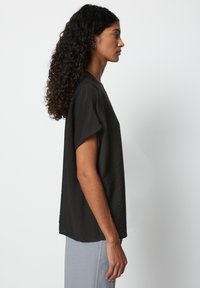 Sort t-shirt med korte ærmer og afslappet pasform, der har en subtil tekstur. Modellen står i profil mod en ensfarvet baggrund.