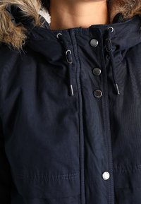 Mørkeblå jakke med pelsforet hætte, sølvknapper, justerbare snore og en blød tekstur. Viser den øverste del, inklusive kraven.