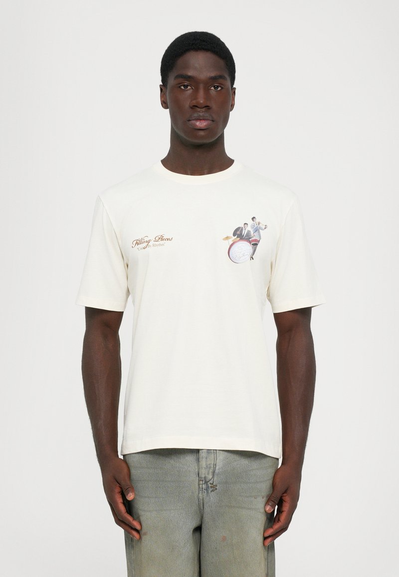 Man wearing white t-shirt with "Filling Pieces" text and small graphic of two musicians playing drum and trumpet on the chest.