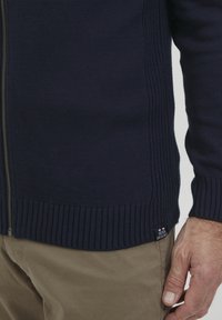 Jūrinis zip-up megztinis su išgaubta tekstūra ir rankovėmis, su šonine etikete. Derinamas su šviesiai smėlio spalvos kelnėmis. Dėmesys medžiagai ir dizainui.