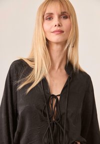 Blusa negra de manga larga con una superficie texturizada, corte holgado y cierre con cordón en el escote. Acentos sutiles realzan el diseño.
